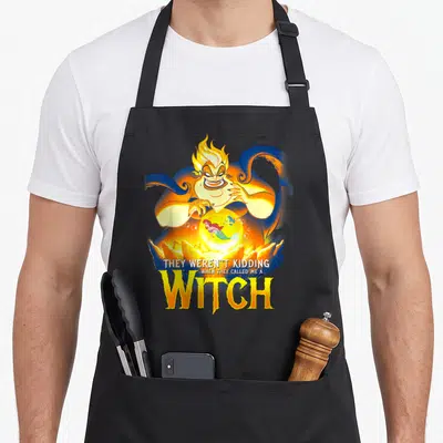 Evil witch apron - Sea witch holding glowing orb with mermaid and fish, stacked yellow WITCH text below — shown on a black cotton canvas apron against a white background