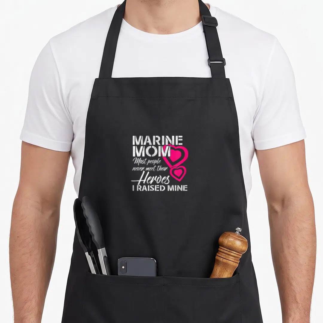 Marine people meet apron - Stacked white text MARINE MOM with two overlapping pink hearts on right side — shown on a black cotton canvas apron against a white background