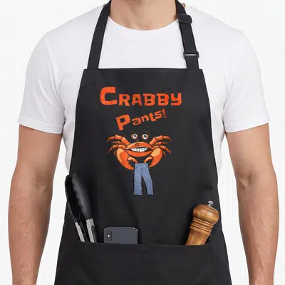 Positive meme apron - Orange text reading Crabby Pants above a cartoon crab wearing blue pants — shown on a black cotton canvas apron against a white background