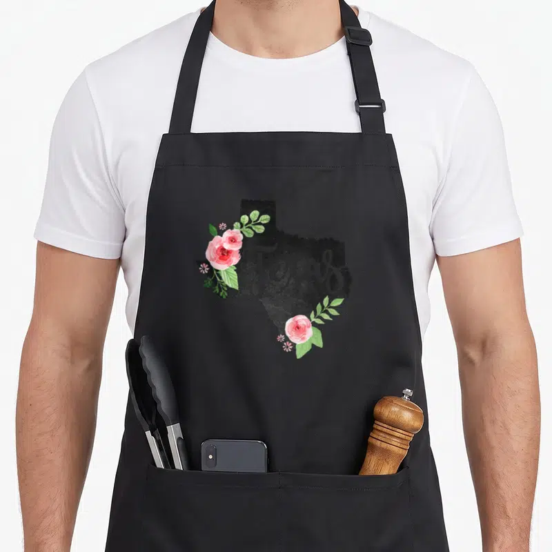 Floral texas apron - Black Texas map silhouette with grey script text and pink watercolor roses on edges — shown on a black cotton canvas apron against a white background