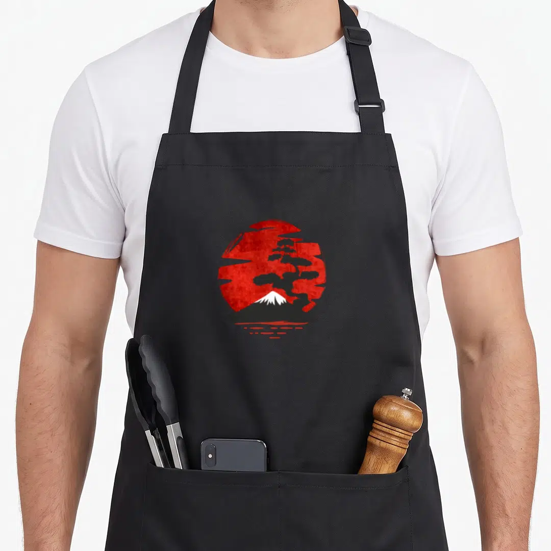 Funny japanese tree apron - Black bonsai and white mountain silhouette against a textured red sun with red water dashes — shown on a black cotton canvas apron against a white background