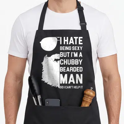 But i love my bearded apron - White silhouette of man with sunglasses and beard next to stacked sarcastic text — shown on a black cotton canvas apron against a white background