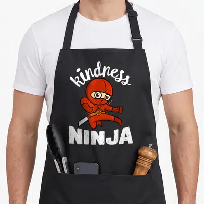 Kind bullying prevention apron - Red cartoon ninja holding sword with kindness arched above and NINJA below in white — shown on a black cotton canvas apron against a white background