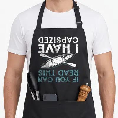 Read canoe girl apron - Upside down kayak and paddle with distressed white and blue text reading CAPSIZED I HAVE READ — shown on a black cotton canvas apron against a white background