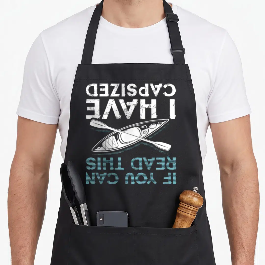 Read canoe girl apron - Upside down kayak and paddle with distressed white and blue text reading CAPSIZED I HAVE READ — shown on a black cotton canvas apron against a white background