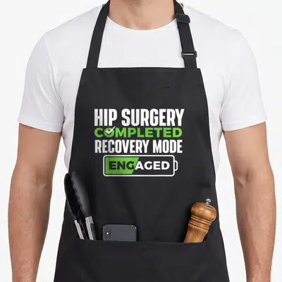 Completed hip apron - Stacked text reading Hip Surgery Completed Recovery Mode Engaged inside green and white battery — shown on a black cotton canvas apron against a white background