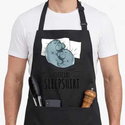 Sleepshirt cow apron - Two blue-grey hippos cuddling on a white pillow under a grey blanket with black text — shown on a black cotton canvas apron against a white background