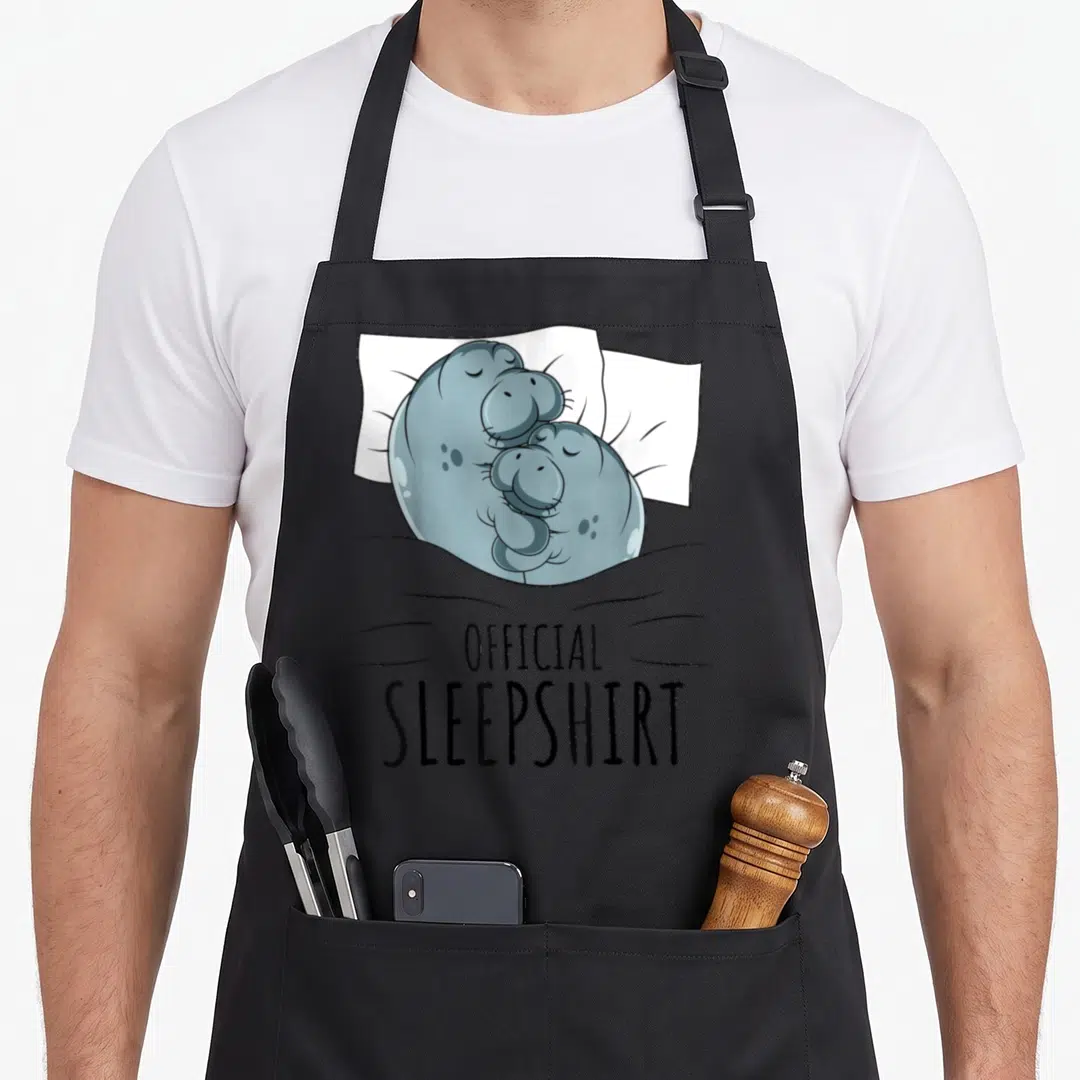 Sleepshirt cow apron - Two blue-grey hippos cuddling on a white pillow under a grey blanket with black text — shown on a black cotton canvas apron against a white background