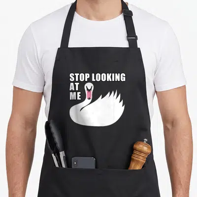 Stop funny swan apron - Cartoon white swan with open pink mouth and text STOP LOOKING AT ME — shown on a black cotton canvas apron against a white background