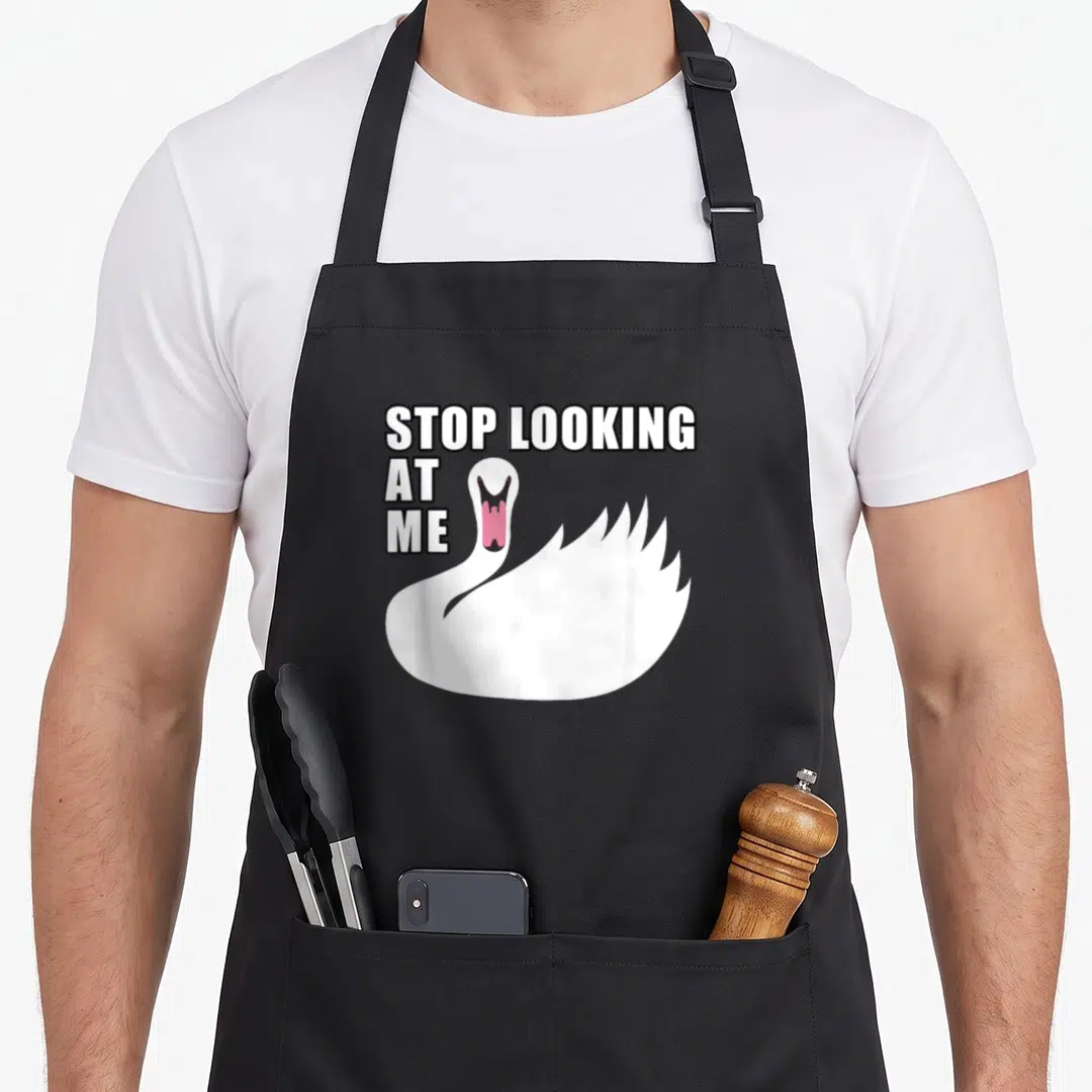 Stop funny swan apron - Cartoon white swan with open pink mouth and text STOP LOOKING AT ME — shown on a black cotton canvas apron against a white background