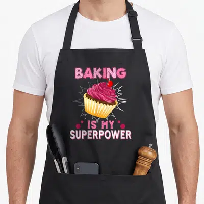 Superpower cupcake apron - Cartoon cupcake with pink frosting and cherry surrounded by Baking Is My Superpower text — shown on a black cotton canvas apron against a white background