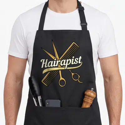 Hairdresser salon hairstylist apron - Gold scissors and comb crossed behind off-white Hairapist script text — shown on a black cotton canvas apron against a white background