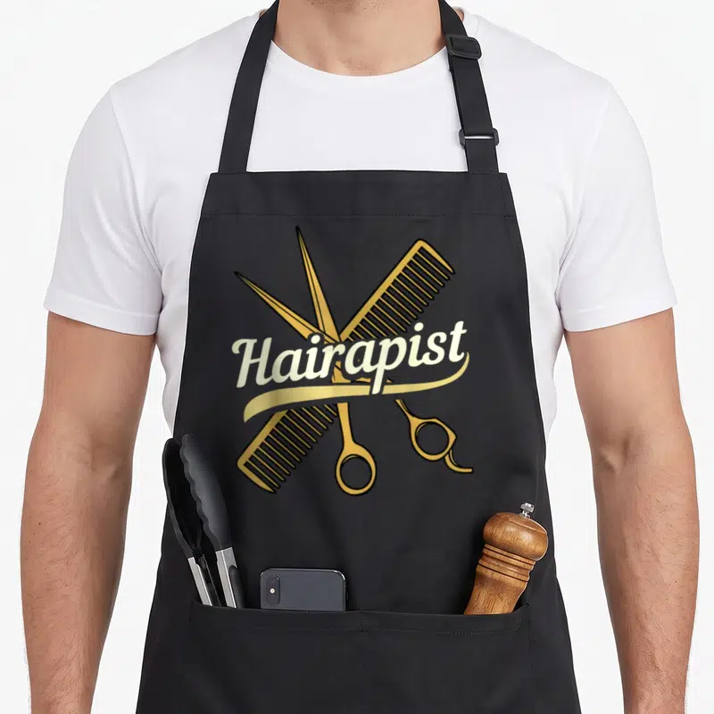 Hairdresser salon hairstylist apron - Gold scissors and comb crossed behind off-white Hairapist script text — shown on a black cotton canvas apron against a white background