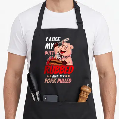 Pulled meat aprons - Cartoon pig holding fork and spatula over flaming meat with stacked pun text — shown on a black cotton canvas apron against a white background