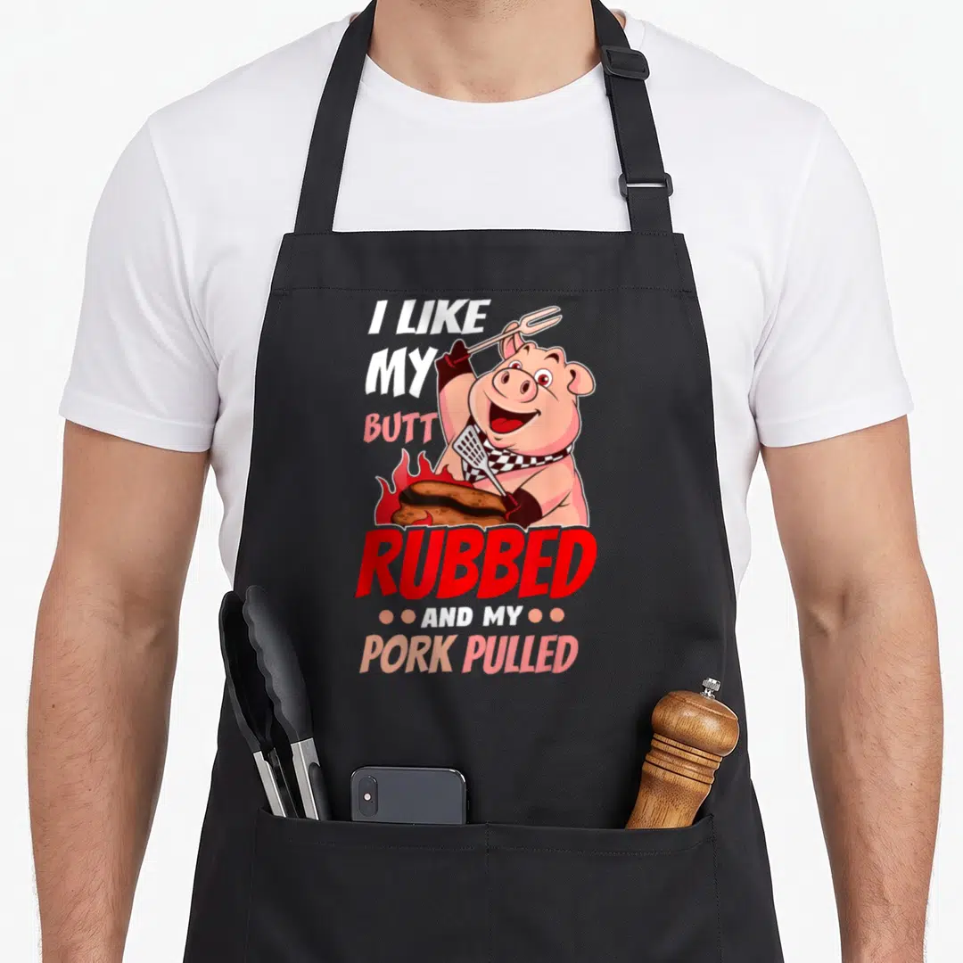Pulled meat aprons - Cartoon pig holding fork and spatula over flaming meat with stacked pun text — shown on a black cotton canvas apron against a white background