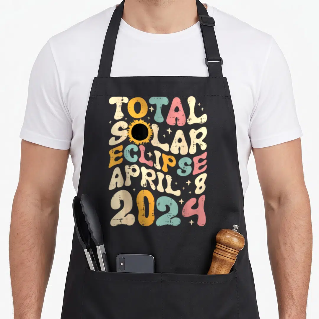 Groovy april apron - Retro colorful typography reading Total Solar Eclipse April 8 2024 with a central eclipse graphic — shown on a black cotton canvas apron against a white background