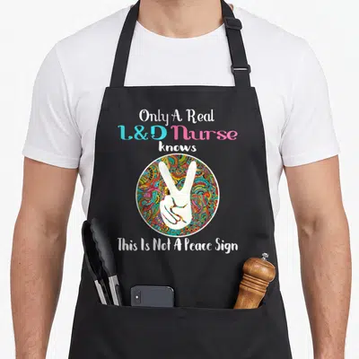 Birth nurse apron - White V-sign hand on multicolored circle with L&D nurse text — shown on a black cotton canvas apron against a white background