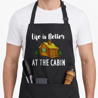Better mountain cabin apron - Cartoon log cabin with yellow roof and white text Life is Better AT THE CABIN — shown on a black cotton canvas apron against a white background