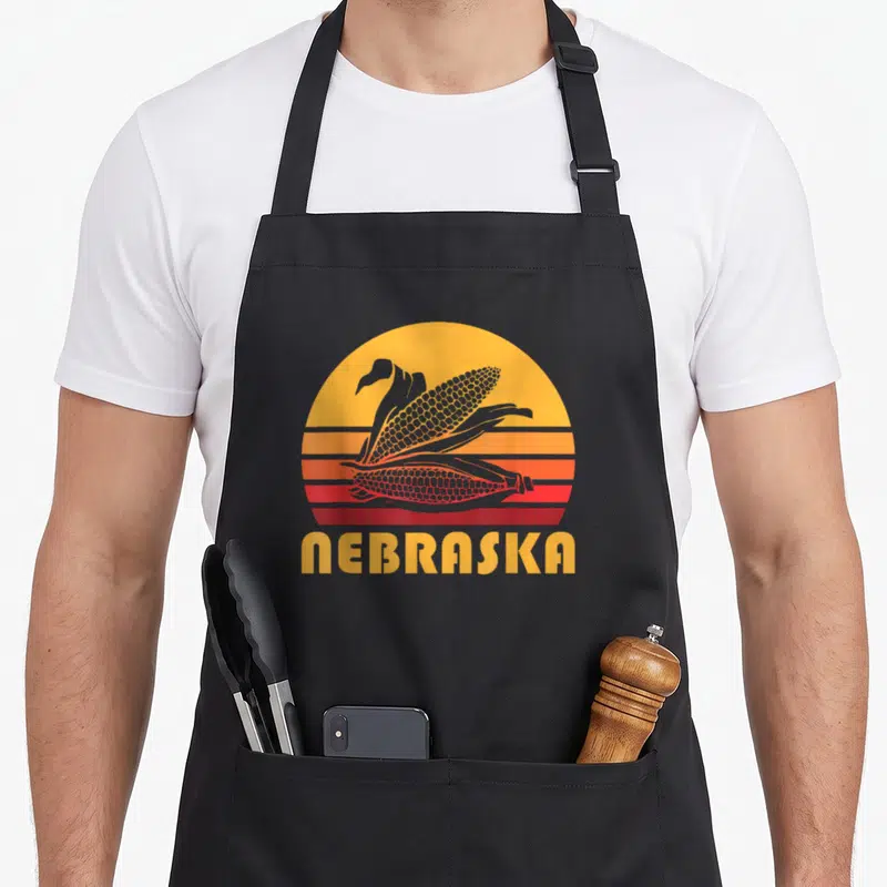 Nebraska farming apron - Two corn ears inside striped sunburst with Nebraska text in retro style — shown on a black cotton canvas apron against a white background