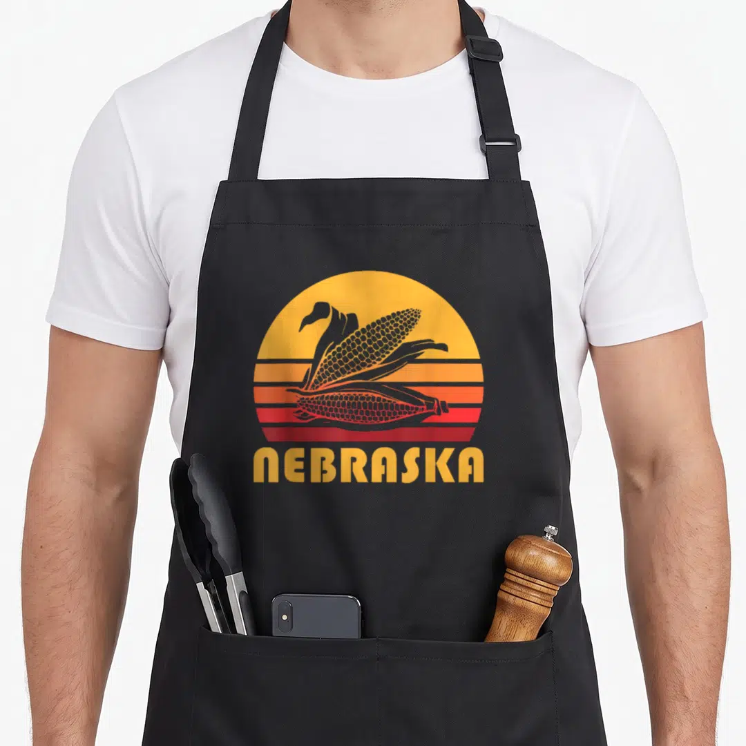 Nebraska farming apron - Two corn ears inside striped sunburst with Nebraska text in retro style — shown on a black cotton canvas apron against a white background