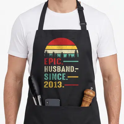 13th wedding apron - EPIC. HUSBAND. SINCE. 2013 text under distressed red yellow beige teal sun and tree silhouette — shown on a black cotton canvas apron against a white background