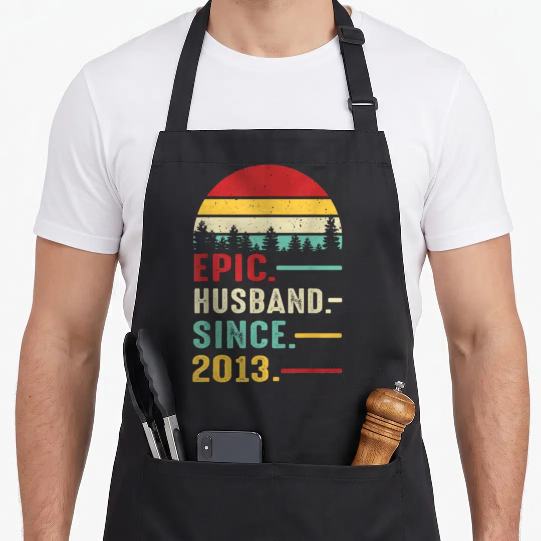 13th wedding apron - EPIC. HUSBAND. SINCE. 2013 text under distressed red yellow beige teal sun and tree silhouette — shown on a black cotton canvas apron against a white background