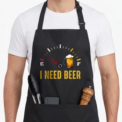 Meter beer brewing apron - Fuel gauge needle at E above beer mug with I NEED BEER text in yellow and red — shown on a black cotton canvas apron against a white background