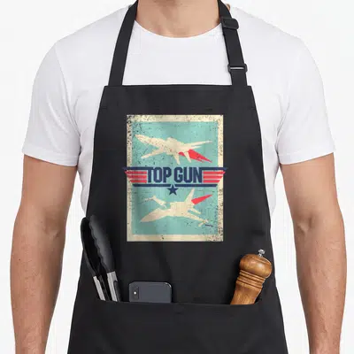 Cutout gun apron - TOP GUN text between two fighter jets with wings and star in cream blue red retro style — shown on a black cotton canvas apron against a white background