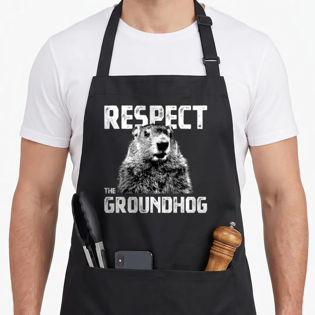Respect hedge hog apron - Distressed white text reading Respect The Groundhog above a grey groundhog illustration — shown on a black cotton canvas apron against a white background