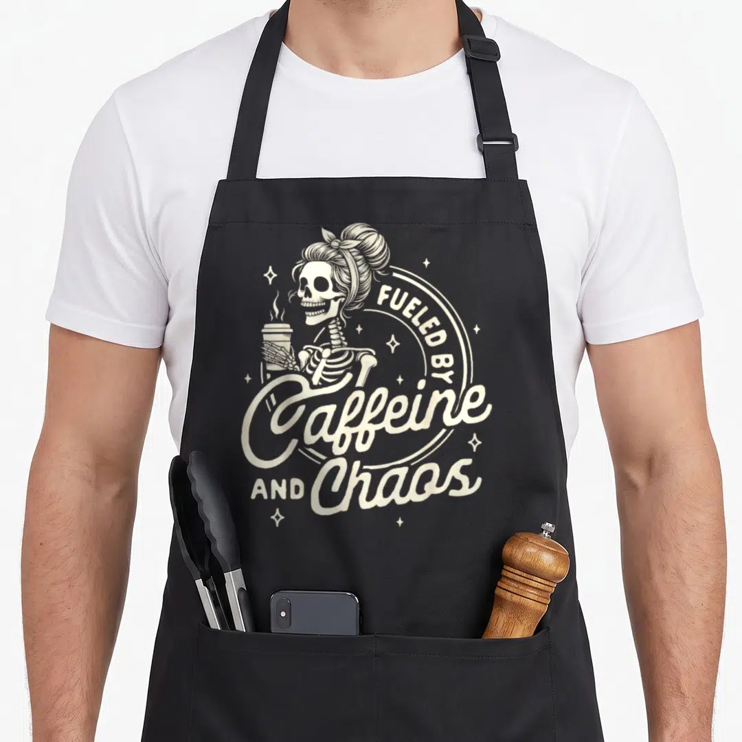 Motherhood coffee apron - Cream skeleton with hair bun holding coffee cup next to Fueled By Caffeine And Chaos text — shown on a black cotton canvas apron against a white background