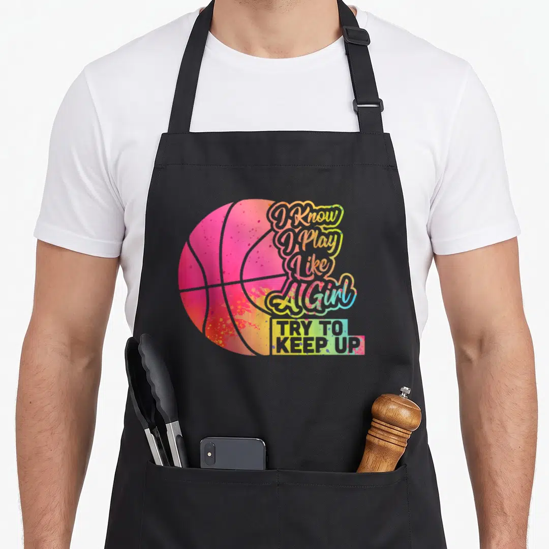 Funny play basketball apron - Rainbow basketball with stacked text I Know I Play Like A Girl Try To Keep Up — shown on a black cotton canvas apron against a white background
