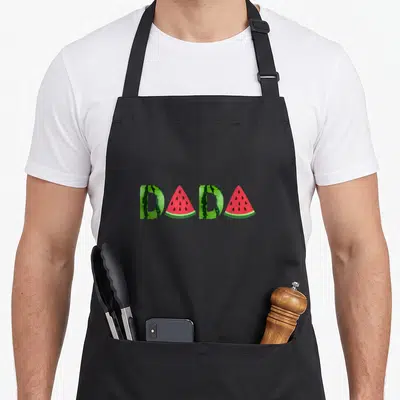 Summer dada watermelon apron - DADA text formed by green watermelon rind and red triangular slices with black seeds — shown on a black cotton canvas apron against a white background
