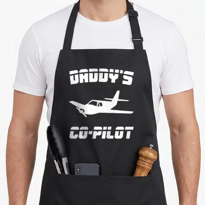 Pilot daddy apron - White airplane silhouette between Daddy's Co-Pilot text on black background — shown on a black cotton canvas apron against a white background