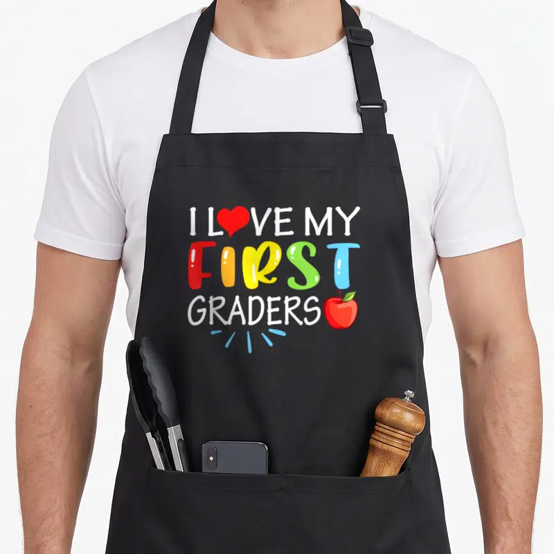 Grader art teacher aprons - I Love My First Graders text with red heart, apple, and blue lines on white and colored — shown on a black cotton canvas apron against a white background