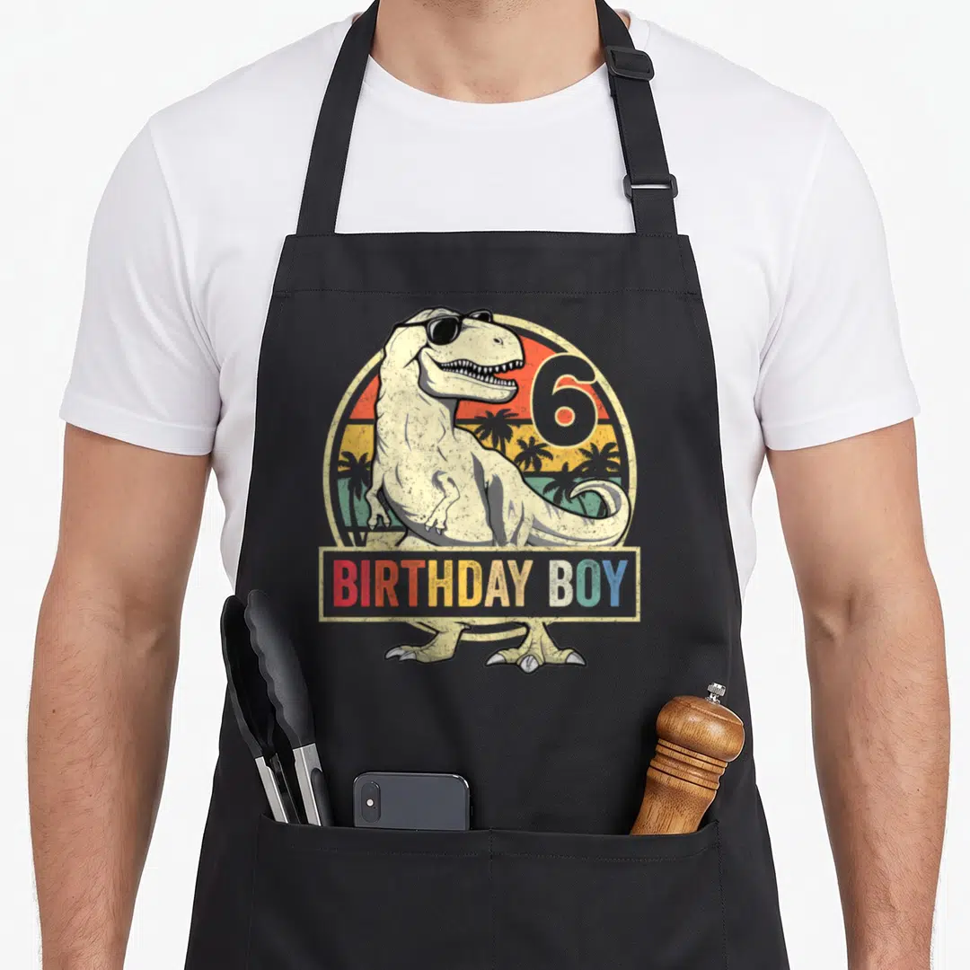 6th birthday t rex apron - T-Rex in sunglasses with 6 BIRTHDAY BOY text on retro sunset background — shown on a black cotton canvas apron against a white background