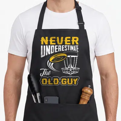 Underestimate disc golf apron - Yellow disc flying toward white basket with distressed NEVER UNDERESTIMATE The OLD GUY — shown on a black cotton canvas apron against a white background