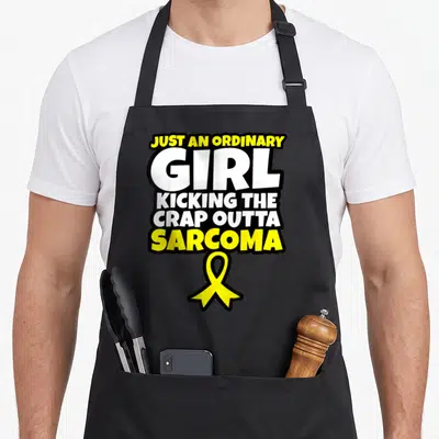 Ordinary ribbon apron - Stacked yellow and white text reading Just an Ordinary Girl Kicking the Crap Outta Sarcoma — shown on a black cotton canvas apron against a white background