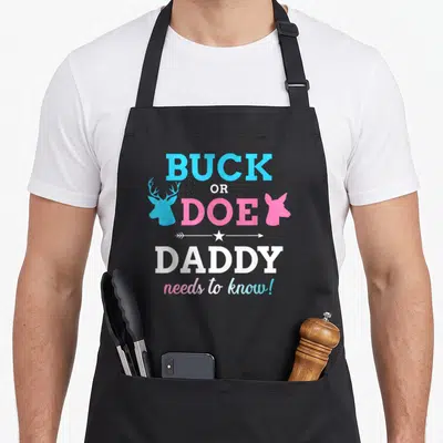 Funny baby daddy apron - Stacked BUCK OR DOE DADDY text with blue buck and pink doe heads and white arrow star — shown on a black cotton canvas apron against a white background