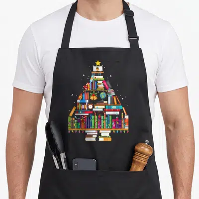 Library christmas tree apron - Stacked books forming a tree shape with star, bells, globe, and text labels like fashion — shown on a black cotton canvas apron against a white background