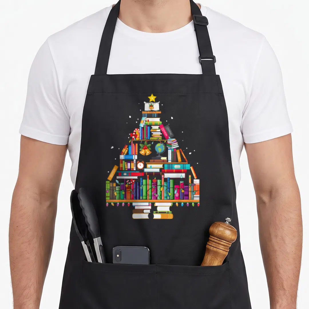 Library christmas tree apron - Stacked books forming a tree shape with star, bells, globe, and text labels like fashion — shown on a black cotton canvas apron against a white background