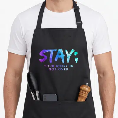 Yet story of the apron - Stacked purple blue and cyan text reading STAY YOUR STORY IS NOT — shown on a black cotton canvas apron against a white background