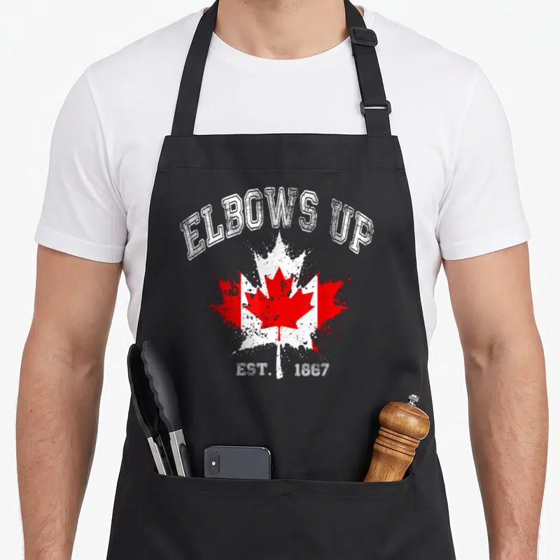 Proud canada aprons - ELBOWS UP text over distressed red maple leaf and flag graphic with EST 1867 below — shown on a black cotton canvas apron against a white background
