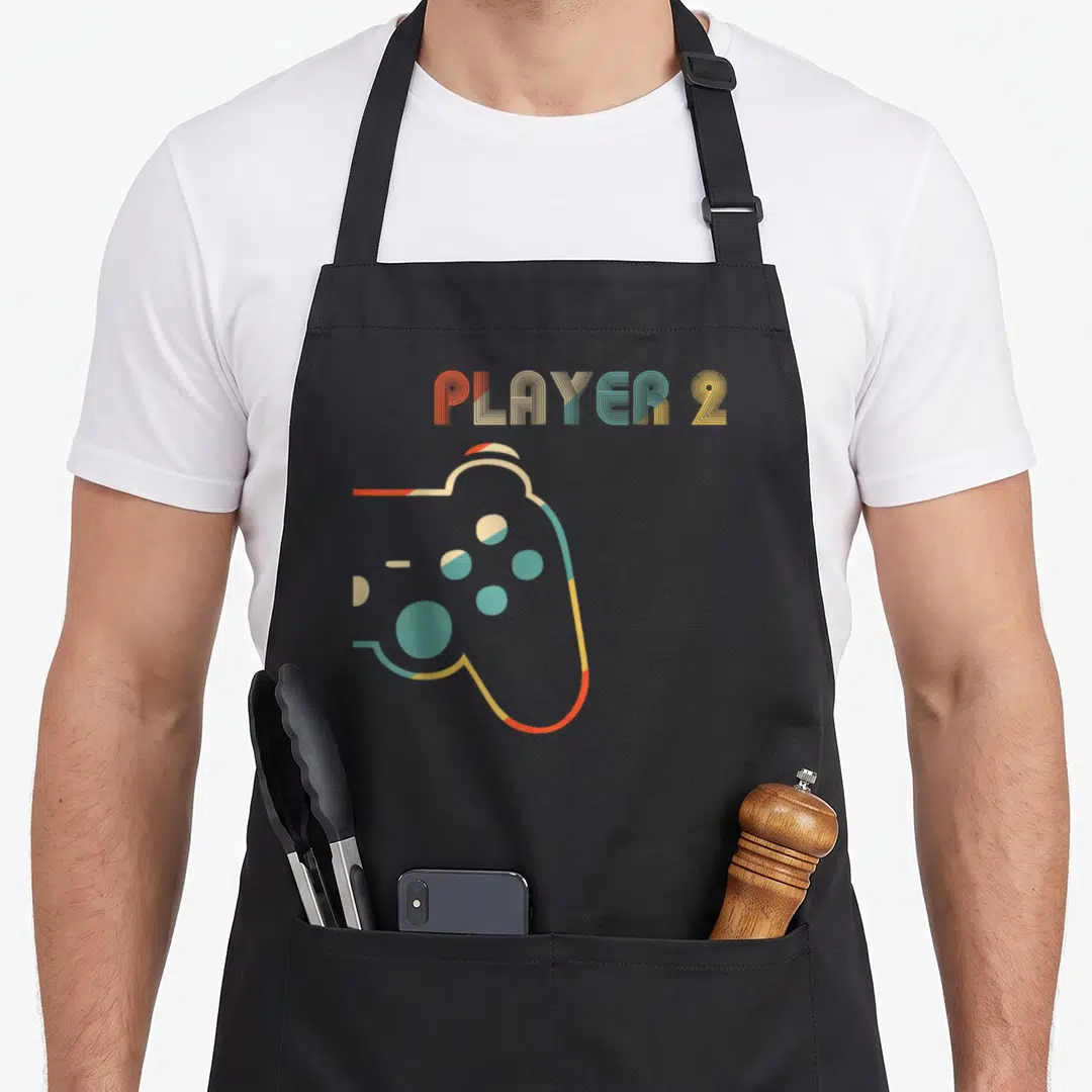 Video couple apron - PLAYER 2 text above orange yellow teal retro game controller graphic — shown on a black cotton canvas apron against a white background