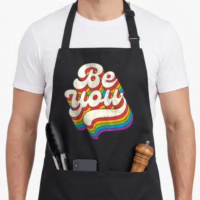Lgbt ally rainbow gay pride apron - Retro 'Be you' text with white letters and multi-colored rainbow shadow extending — shown on a black cotton canvas apron against a white background