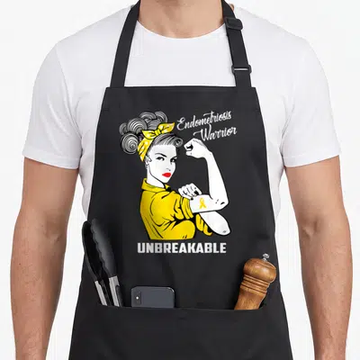 Warrior endometriosis awareness apron - Cartoon woman flexing arm with yellow ribbon and text Endometriosis Warrior — shown on a black cotton canvas apron against a white background