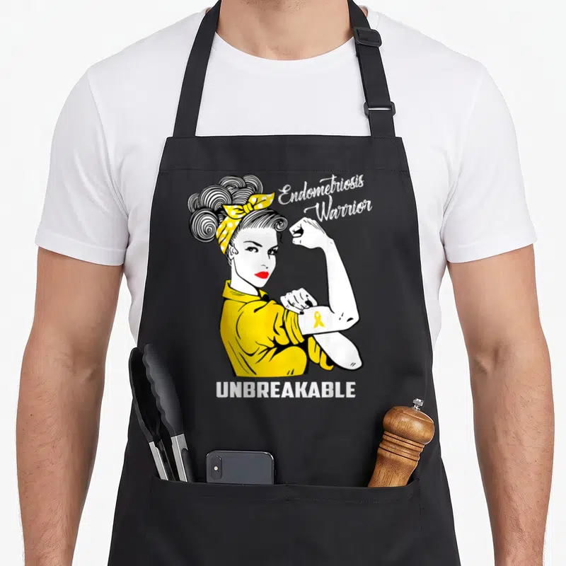 Warrior endometriosis awareness apron - Cartoon woman flexing arm with yellow ribbon and text Endometriosis Warrior — shown on a black cotton canvas apron against a white background