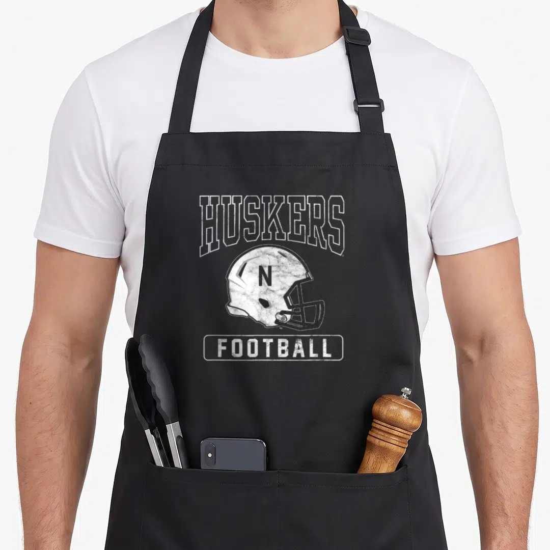 Nebraska football apron - White distressed football helmet with HUSKERS text and N logo — shown on a black cotton canvas apron against a white background