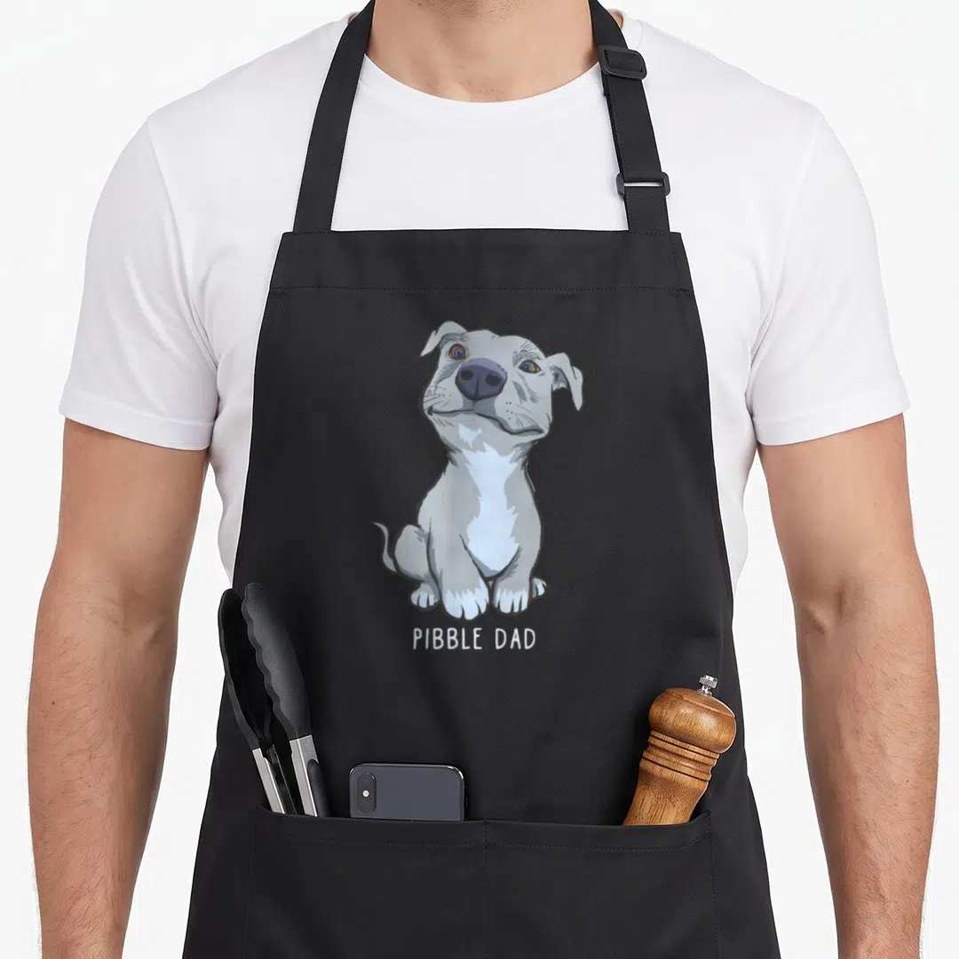 Pibble father day apron - Grey cartoon dog sitting above white PIBBLE DAD text on black fabric — shown on a black cotton canvas apron against a white background