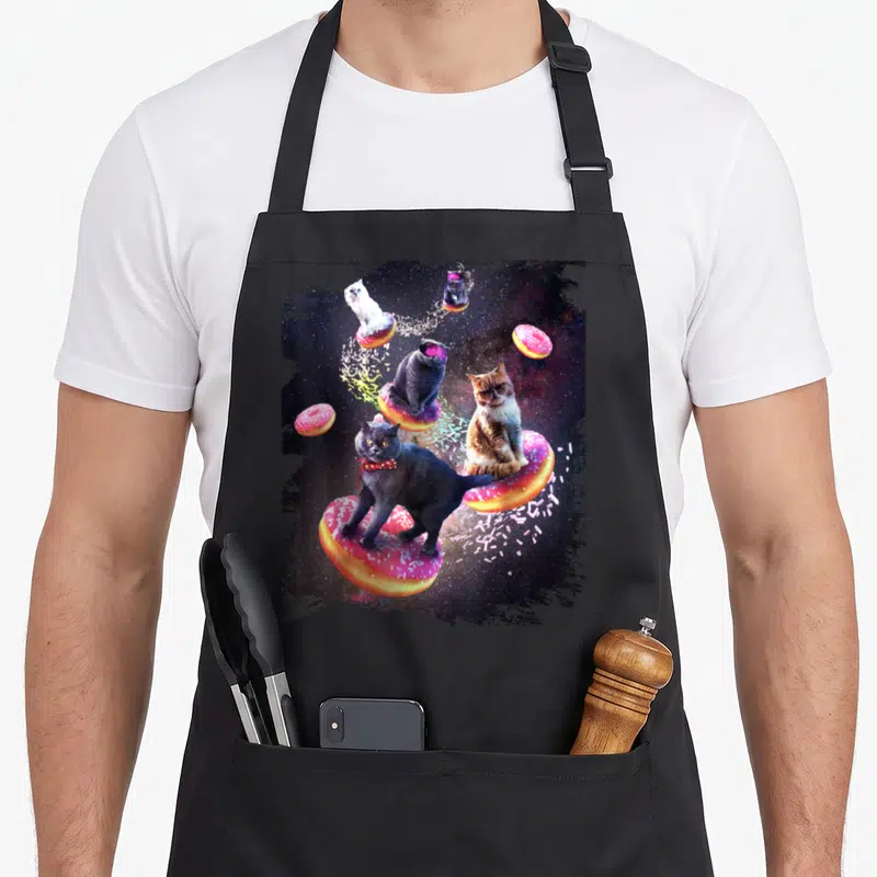 Donut space cat apron - Four cats on pink frosted donuts floating in space with scattered sprinkles — shown on a black cotton canvas apron against a white background