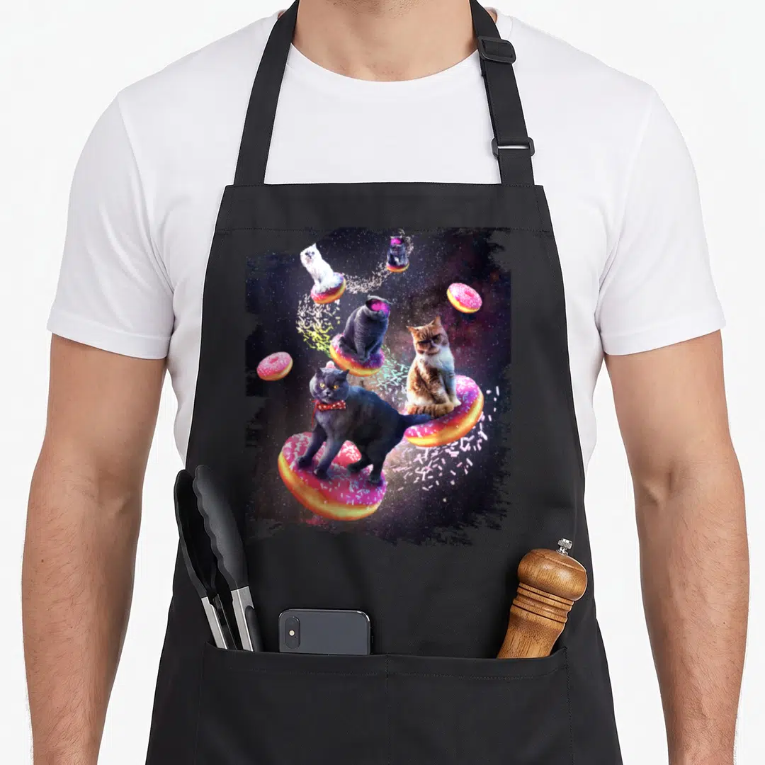 Donut space cat apron - Four cats on pink frosted donuts floating in space with scattered sprinkles — shown on a black cotton canvas apron against a white background